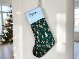 Personalized Christmas stocking hanging on a white wall, with a white cuff embroidered 'Fynn' and a green festive pattern featuring reindeer and snowflakes