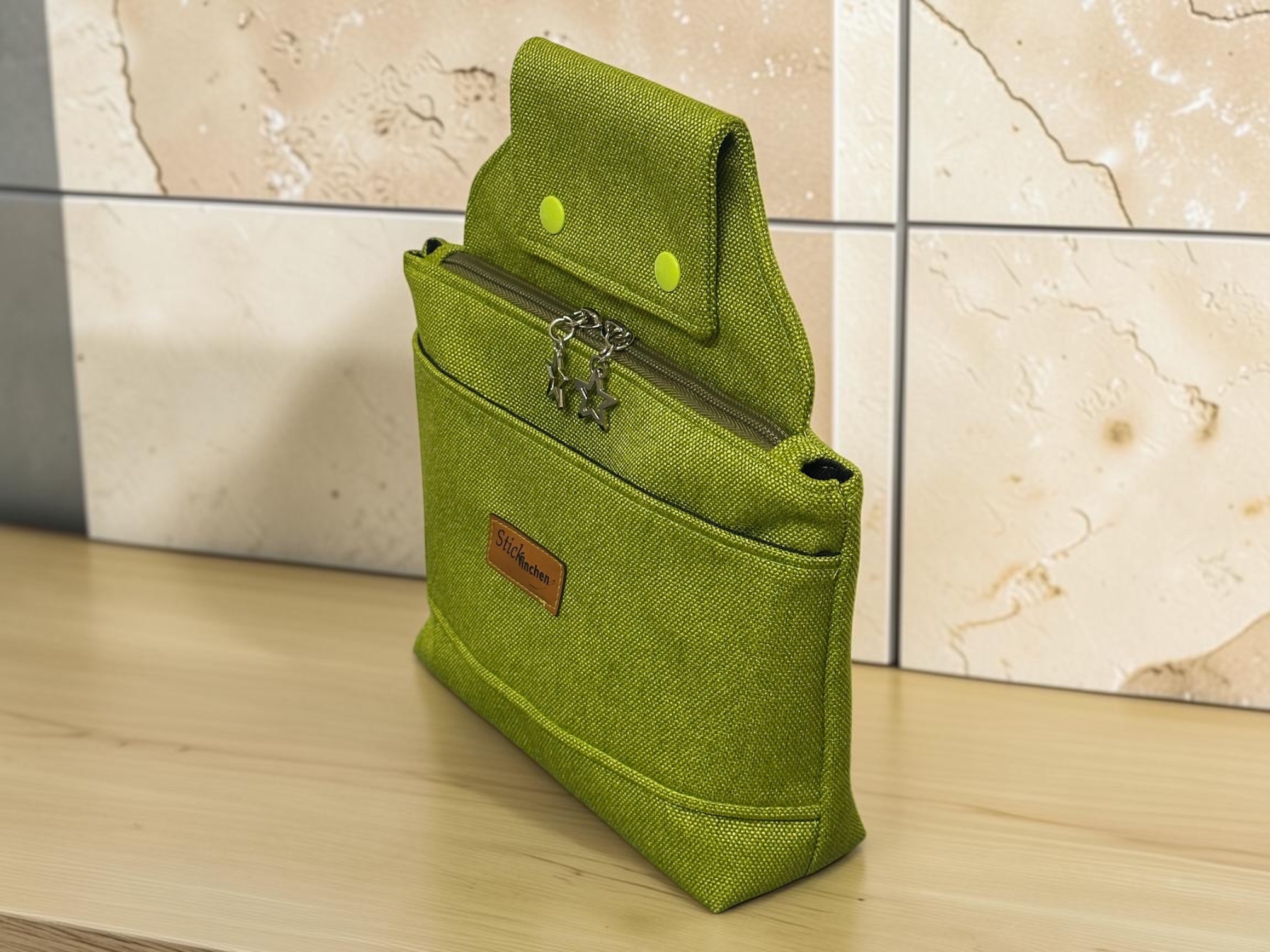 Green textured fabric backpack standing upright on a light wooden table with a zipper and star-shaped charms hanging from the zipper pullages.