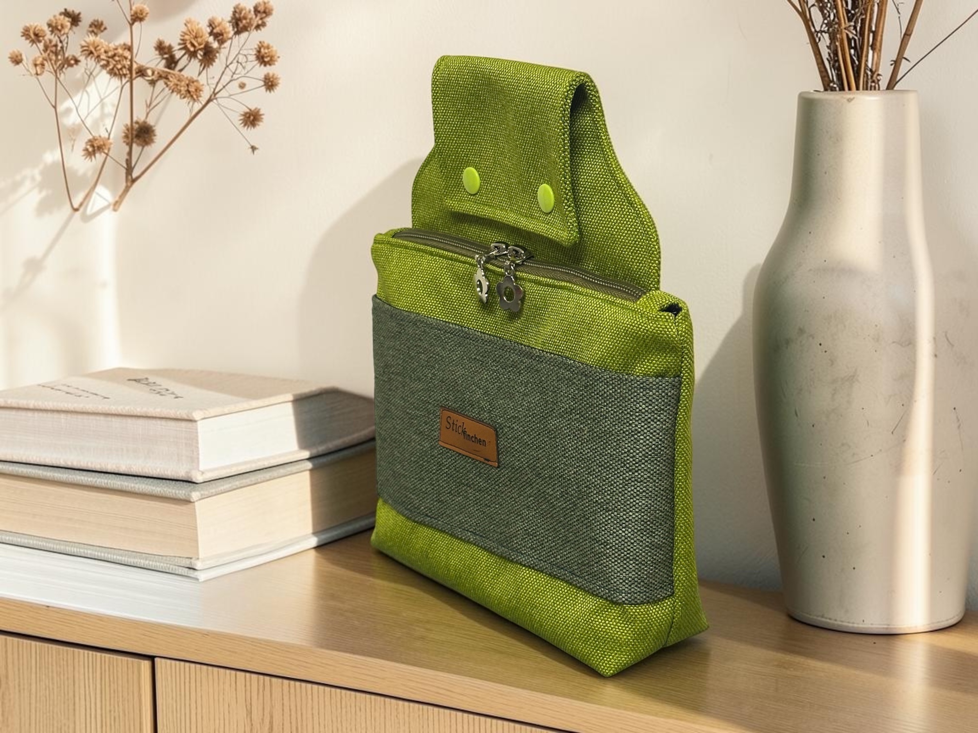 Green two-tone fabric backpack stands on a wooden shelf beside a vase of dried flowers and a book box.