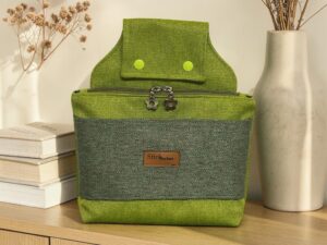 Green fabric backpack on a wooden shelf, front view with zipper pockets and flower-shaped zipper pulls, flanked by dried flowers and a ceramic vase.