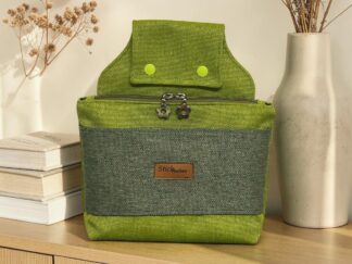 Green fabric backpack on a wooden shelf, front view with zipper pockets and flower-shaped zipper pulls, flanked by dried flowers and a ceramic vase.