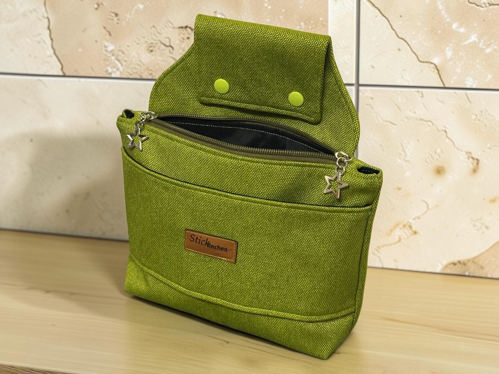 Green textured backpack with star-shaped zipper pulls resting on a light wooden surface, against a tiled wall; small brown brand label on the front pocket.