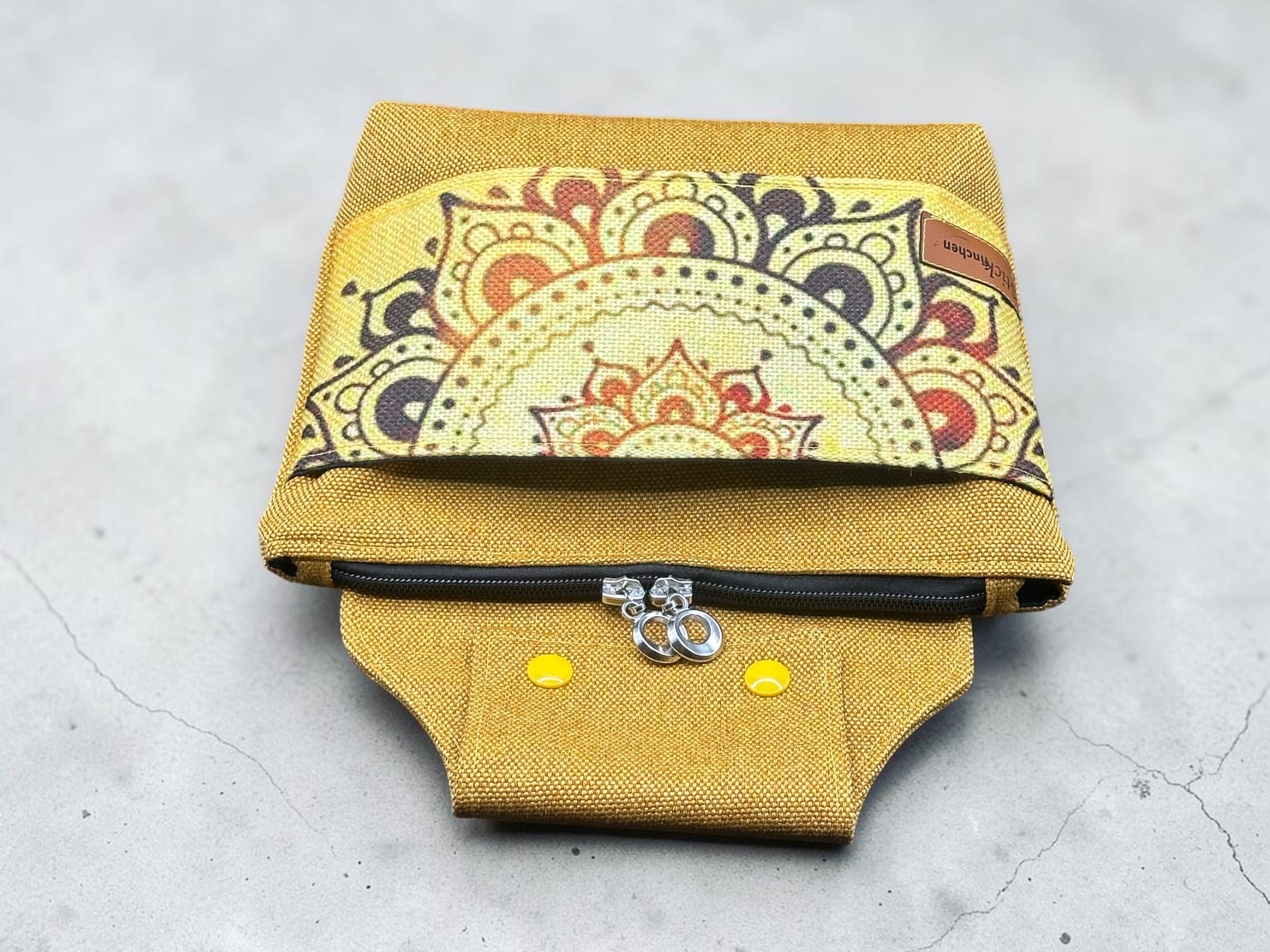Mustard-yellow fabric pouch with a colorful mandala-patterned flap and a visible zipper along the bottom edge.
