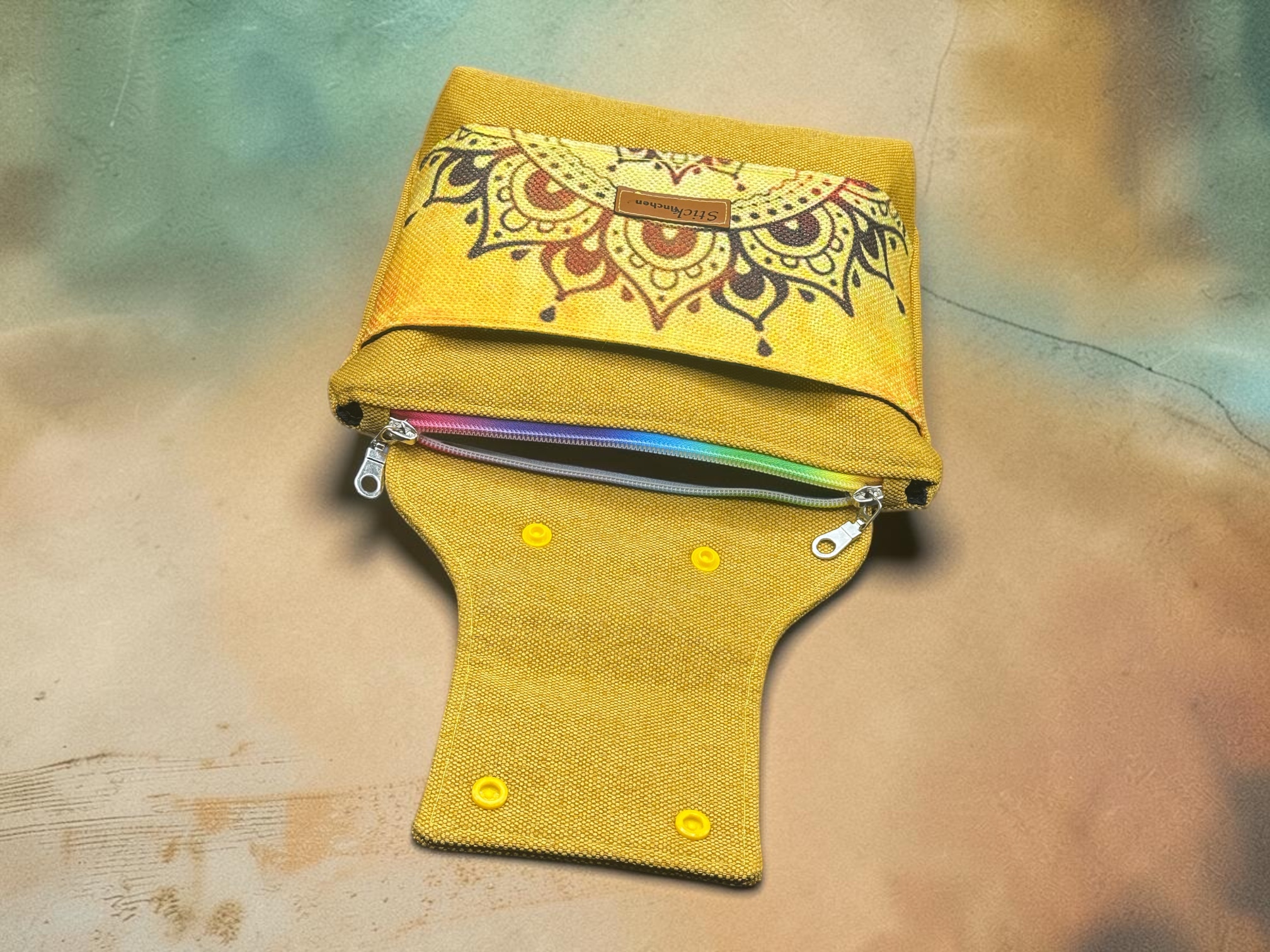Yellow fabric wallet featuring a decorative mandala pattern and a rainbow-colored zipper, set on a textured beige-green background.