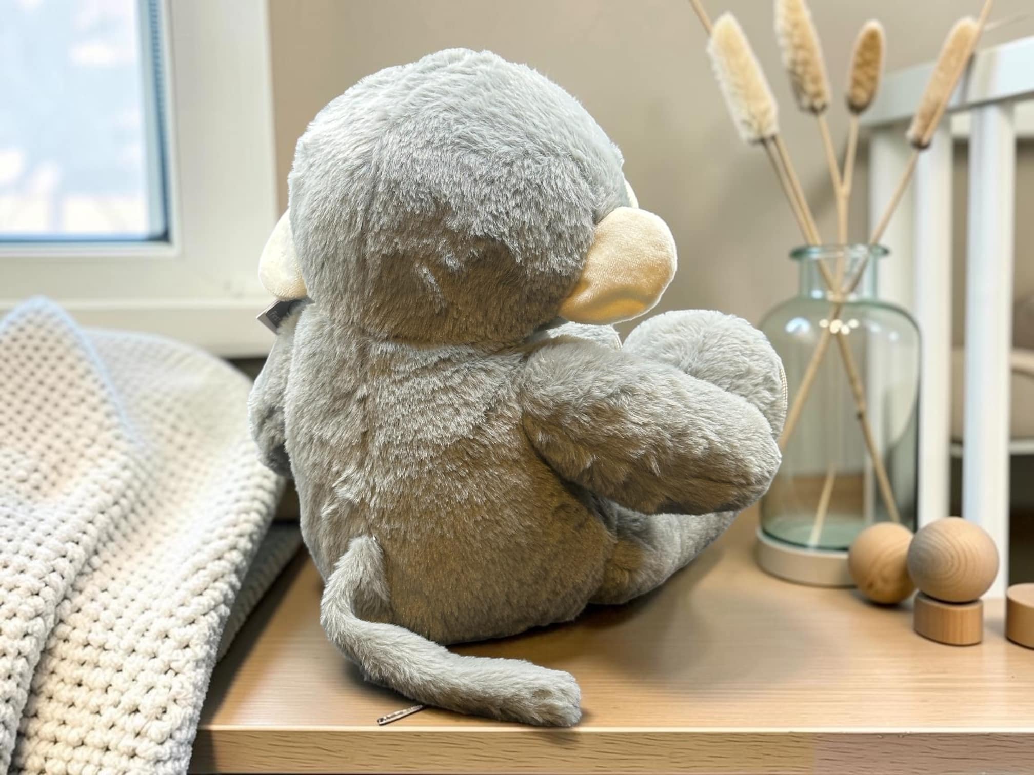 Soft gray plush koala toy resting on a wooden shelf beside a knitted blanket and decorative vase with dried reeds.