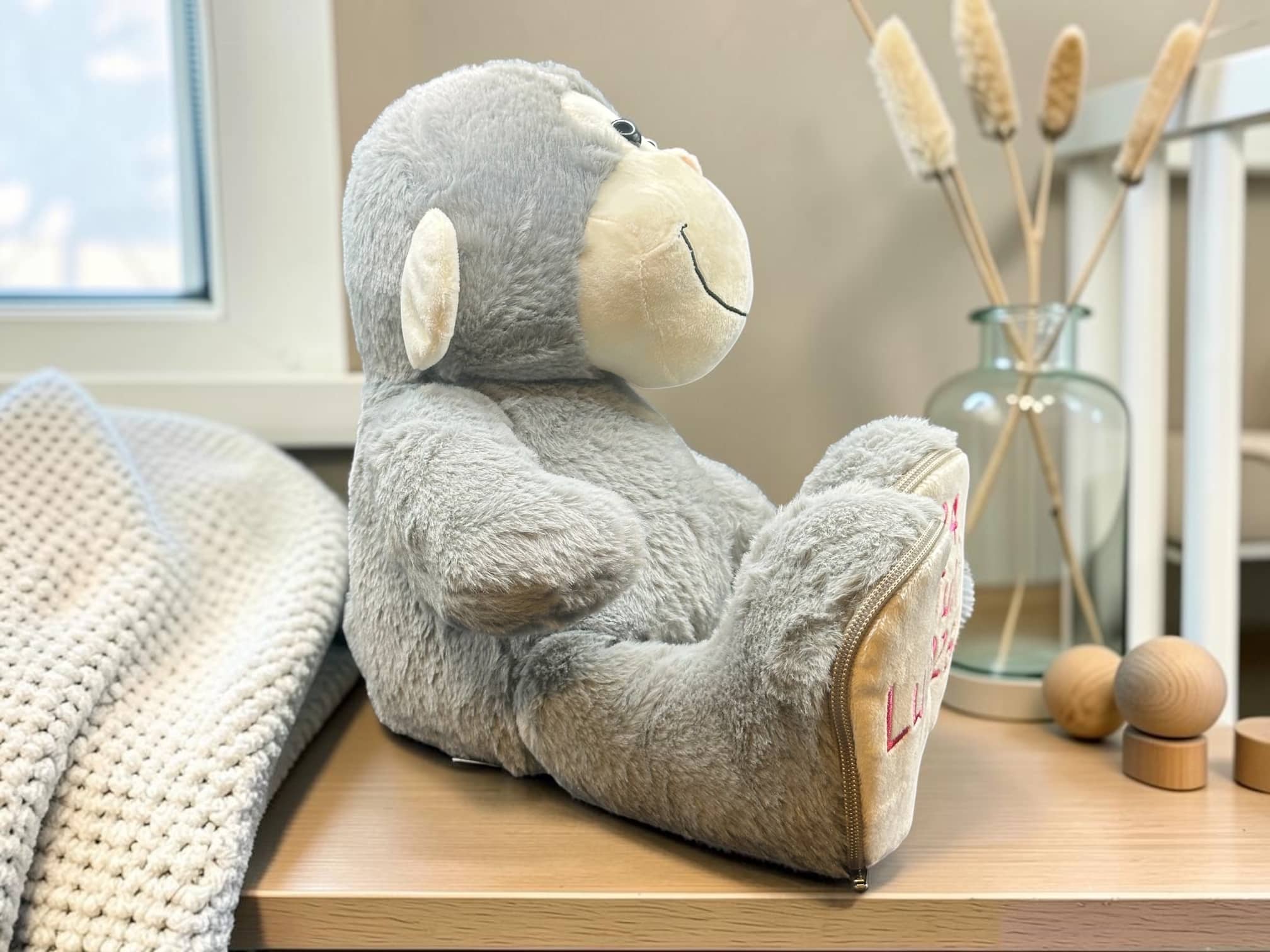 Gray plush teddy bear sitting on a wooden table beside a chunky knit blanket, with a glass vase and dried reeds in the background.