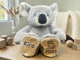 Gray plush koala with beige paw pads sits upright on a wooden table, feet showing the year 2026. Background includes wooden blocks and a cozy room.